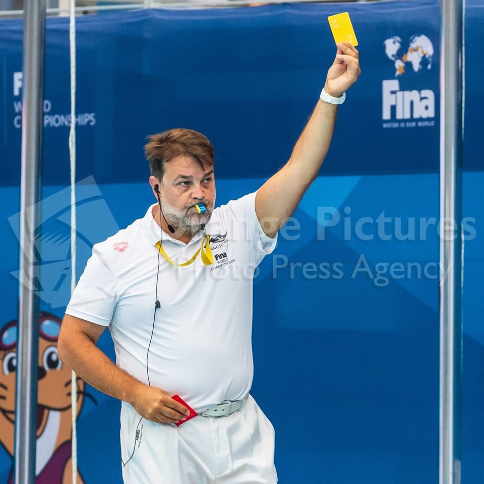 Jaume Teixidó, vestido de blanco con el uniforme FINA levanta el brazo izquierdo para mostrarle la tarjeta amarilla a un jugador. Foto: Orange Pictures